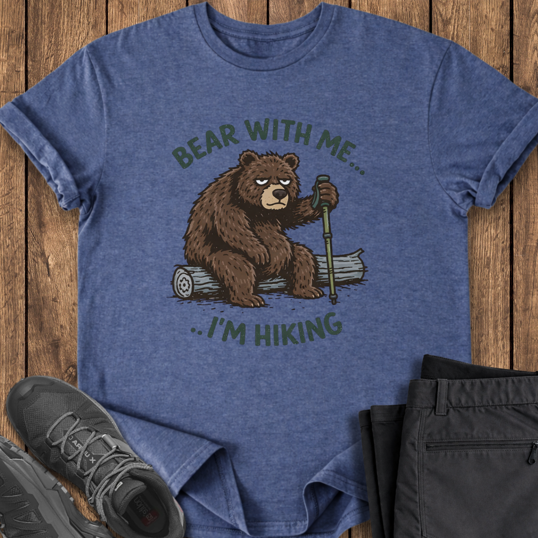 Blue t-shirt with bear graphic and text 'Bear with me... I'm hiking' on a wooden surface with shoes and shorts. grumpy bear hiking funny t shirt