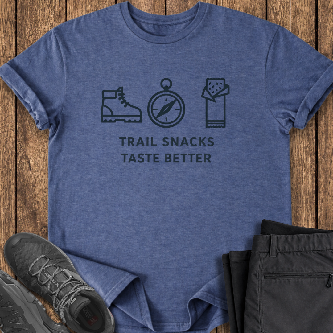 Blue t-shirt with hiking-themed design on a wooden surface with shoes and pants. trail snacks tase better