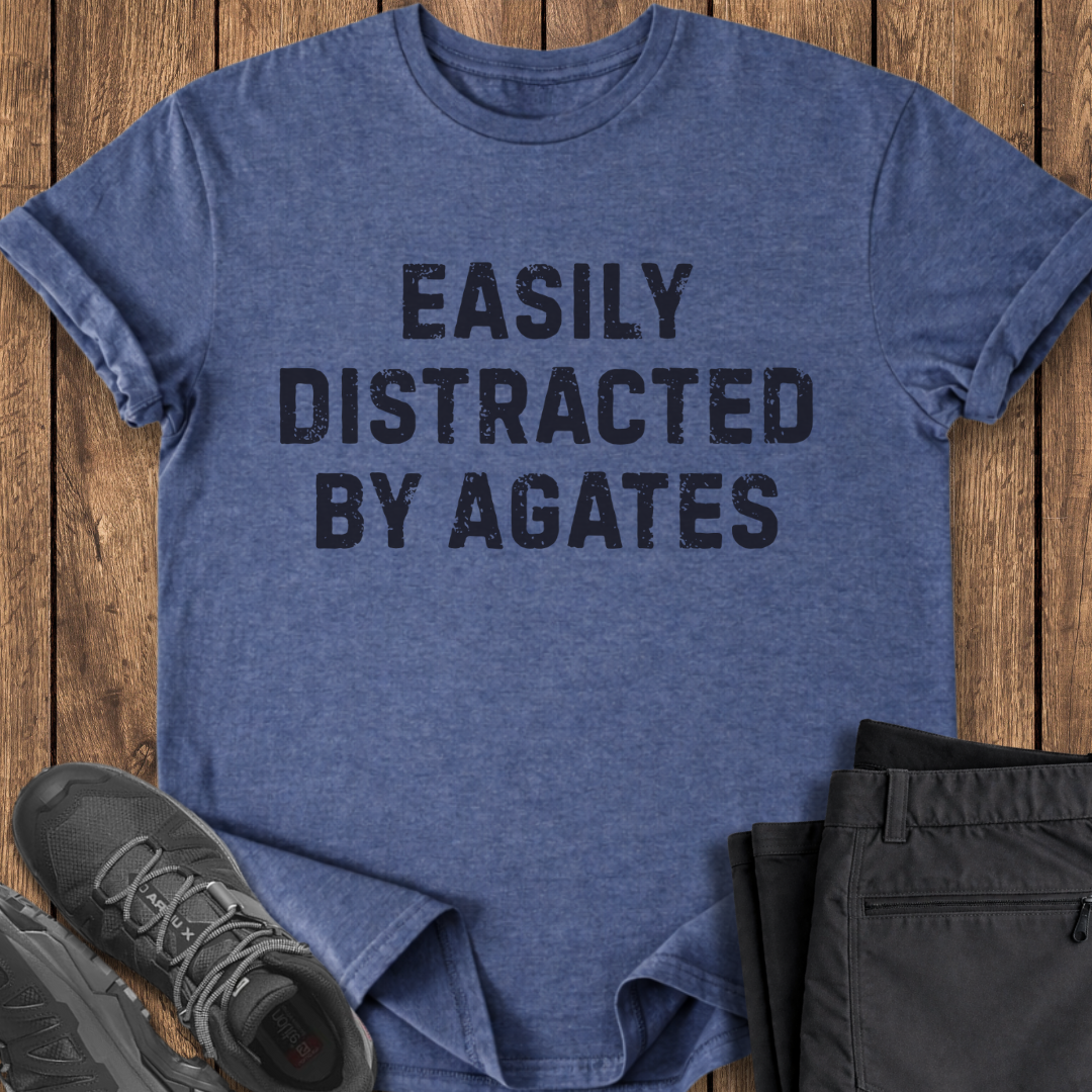 Blue t-shirt with 'EASILY DISTRACTED BY AGATES' text on a wooden surface with shoes and pants.