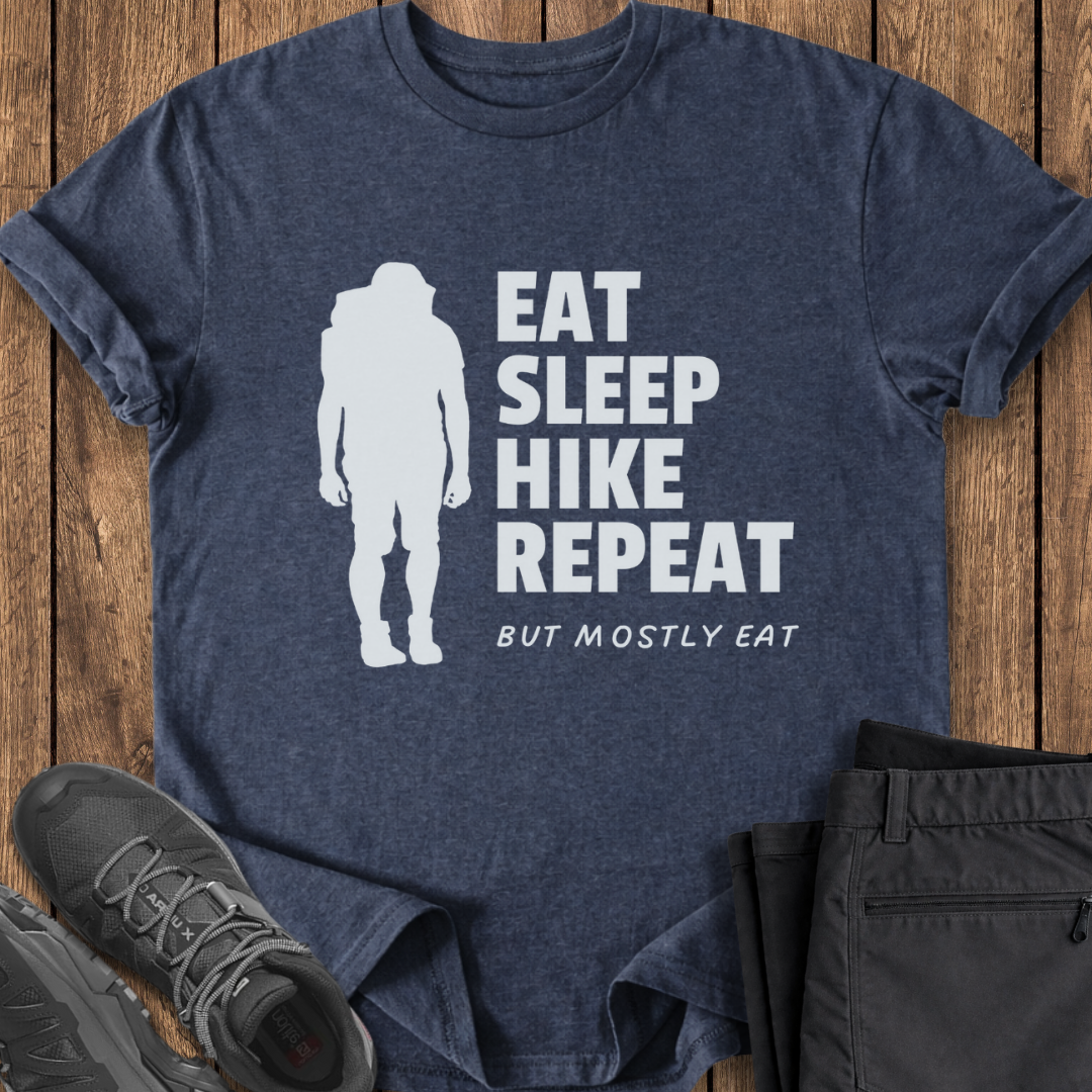 Blue t-shirt with 'Eat Sleep Hike Repeat' text and hiking silhouette on a wooden surface with shoes and pants. funny hiking shirt