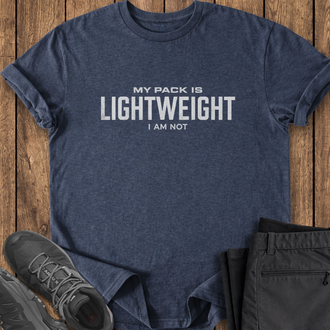 Navy t-shirt with 'My pack is lightweight, I am not' text on a wooden surface with black pants and shoes. funny backpacking t shirt