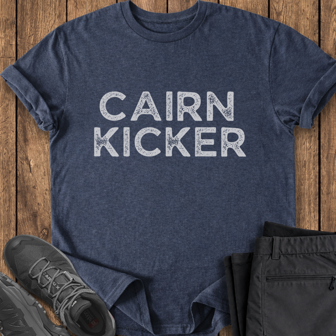 Navy t-shirt with 'CAIRN KICKER' text on a wooden surface with shoes and pants. hiking shirt