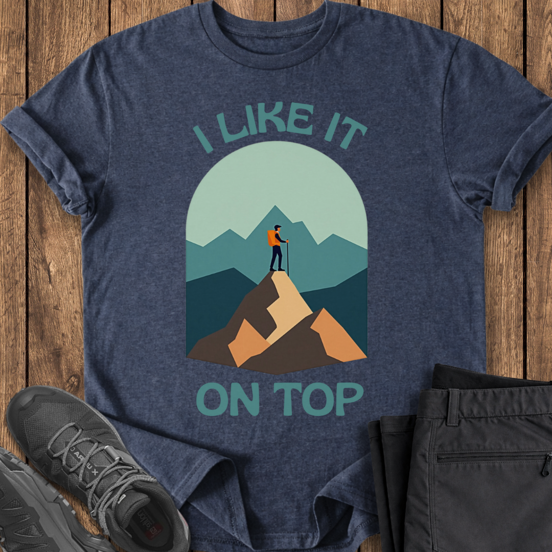 Blue t-shirt with mountain graphic and text 'I Like It On Top' on a wooden surface with shoes and pants.
