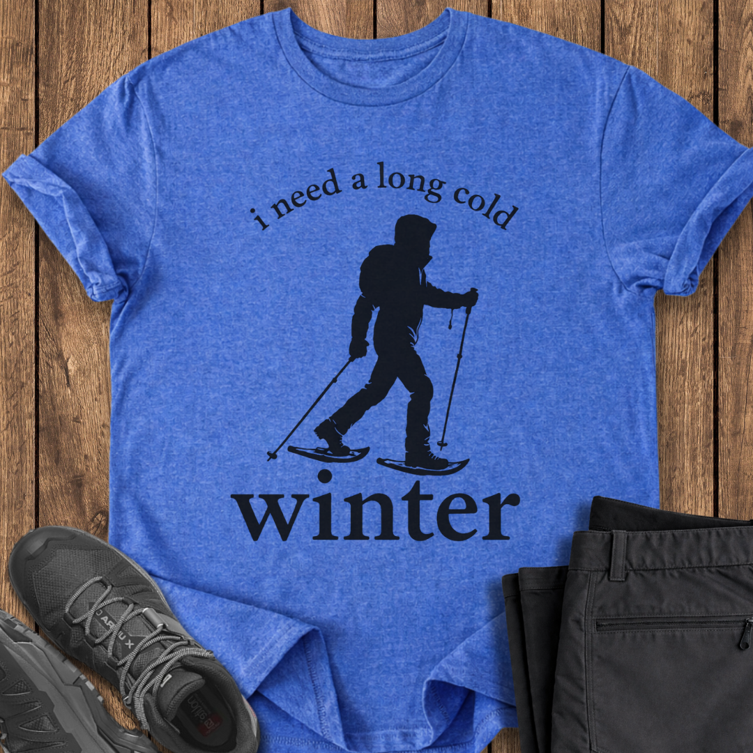 Blue t-shirt with skiing graphic and text on a wooden surface, with shoes and pants in the foreground. winter snowshoeing t shirt