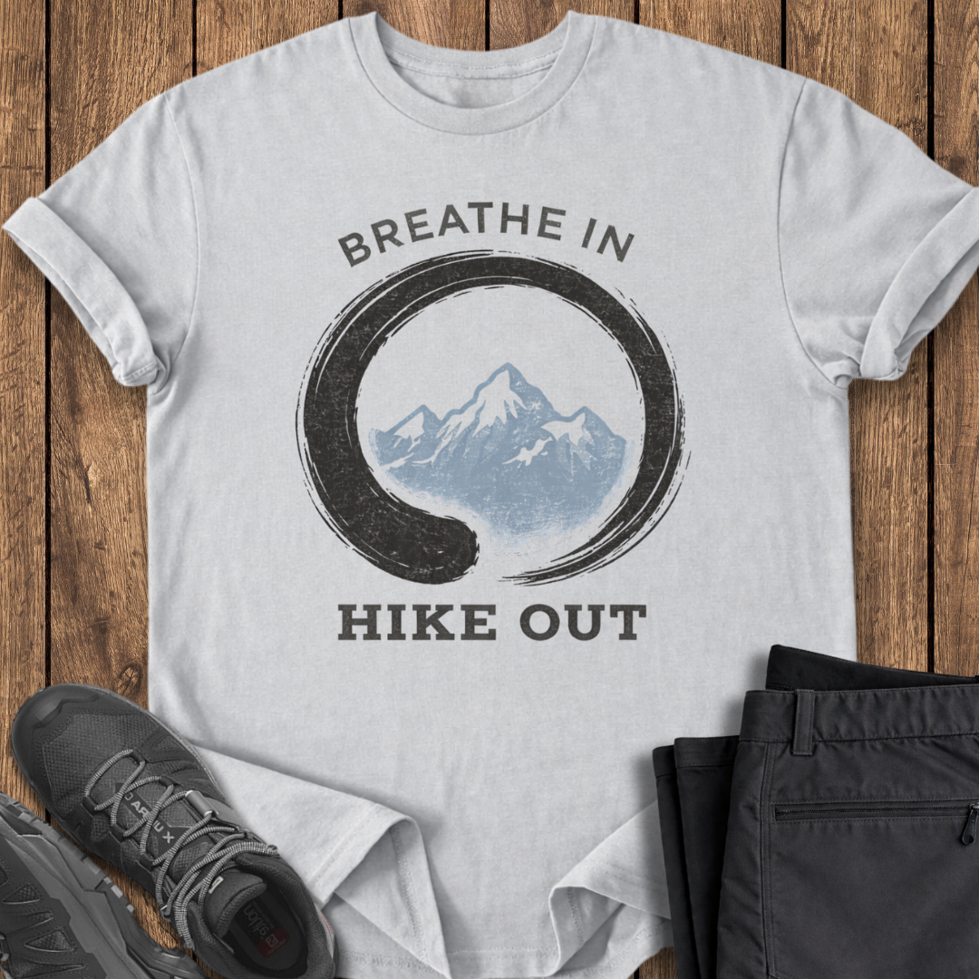 grey t-shirt with mountain graphic and 'Breathe In Hike Out' text on a wooden surface with hiking shoes and pants. mindful hiking t shirt