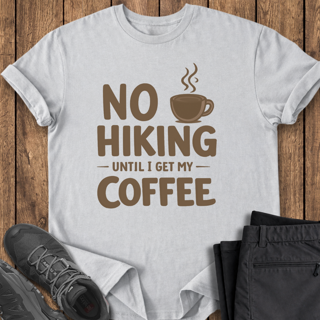 White t-shirt with 'No Hiking Until I Get My Coffee' text and graphic on a wooden surface with shoes and pants. funny hiking t shirt