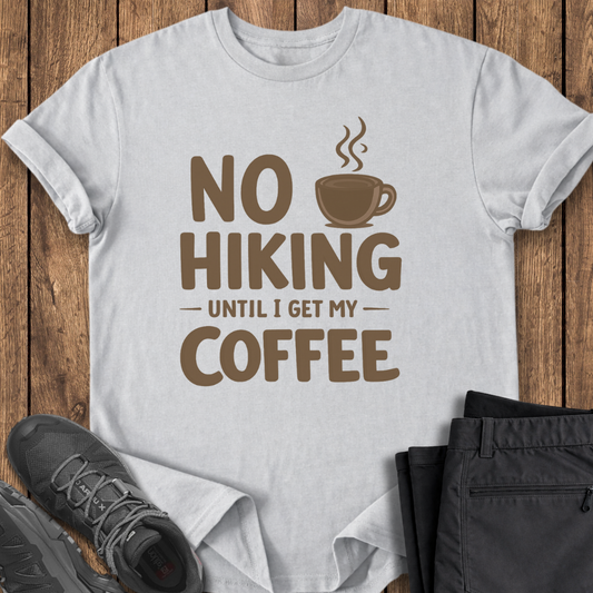 White t-shirt with 'No Hiking Until I Get My Coffee' text and graphic on a wooden surface with shoes and pants. funny hiking t shirt