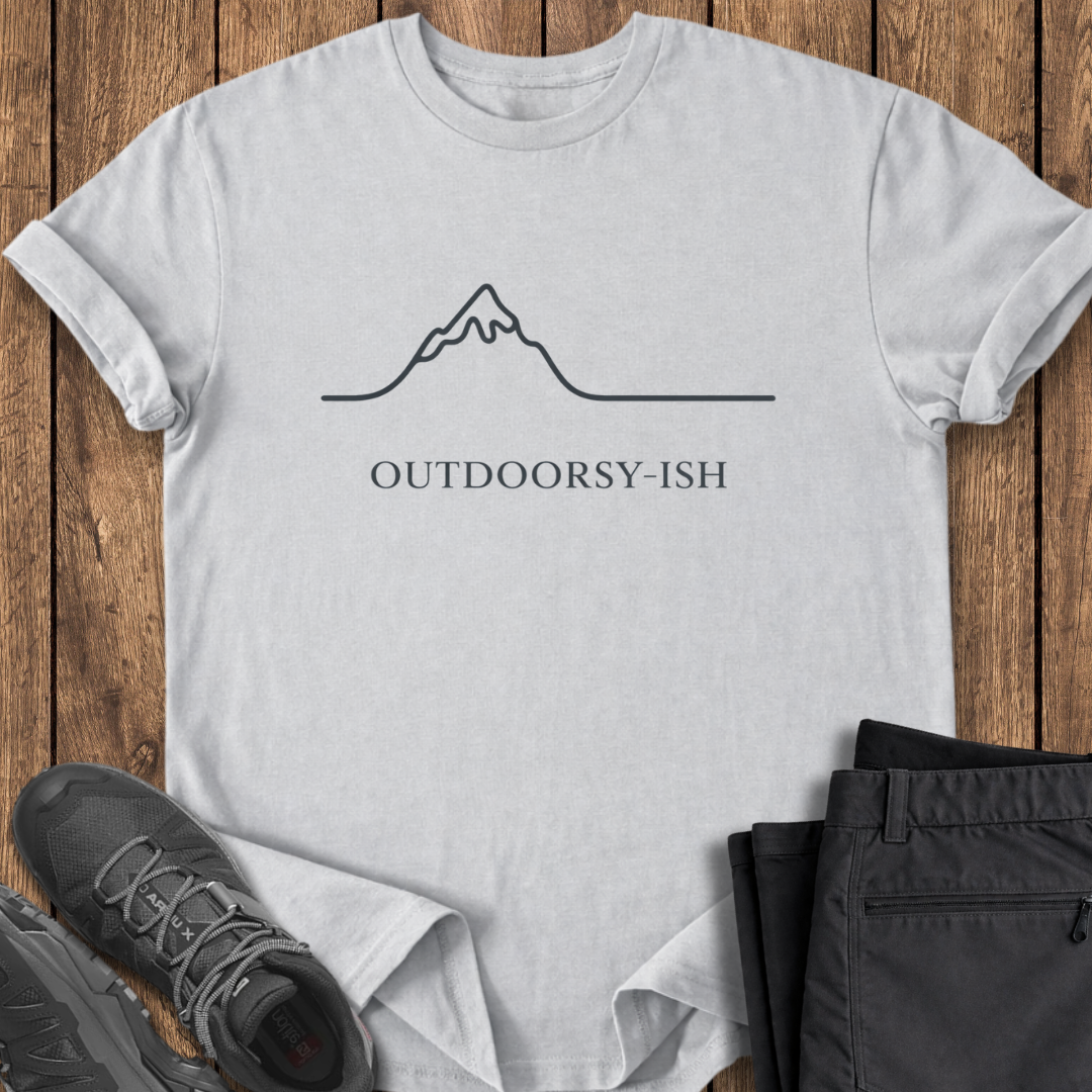White t-shirt with mountain graphic and 'OUTDOORSY-ISH' text on a wooden surface with shoes and pants. funny minimalist mountain shirt