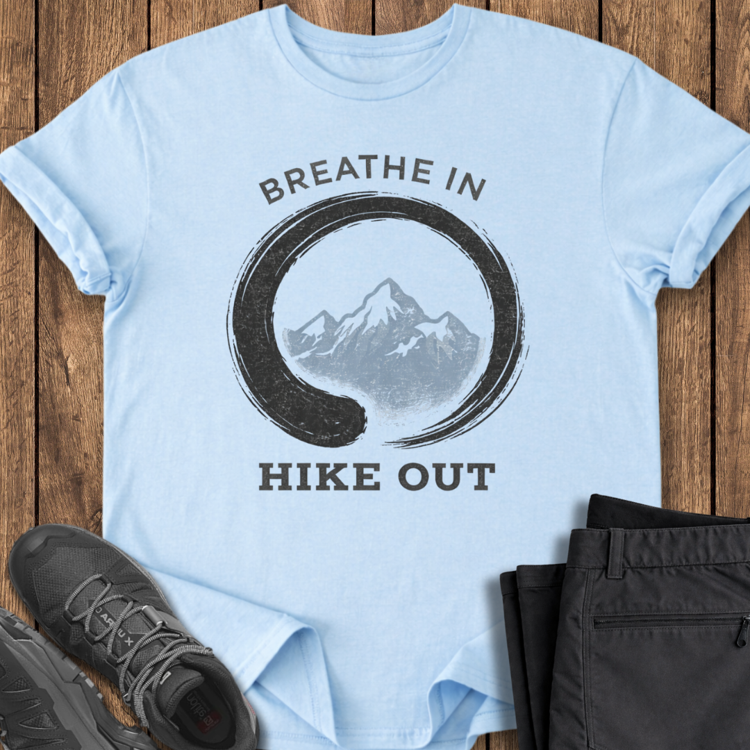 Light blue t-shirt with mountain graphic and 'Breathe In Hike Out' text on a wooden surface with hiking shoes and pants. mindful t shirt
