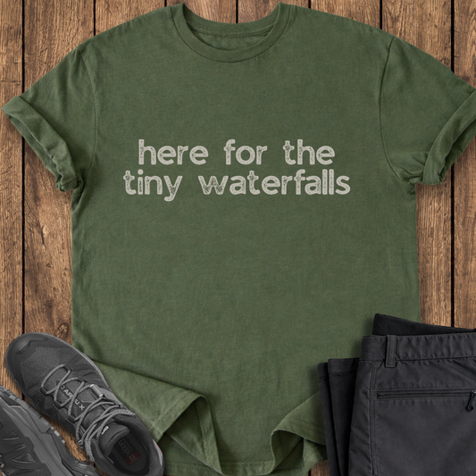 Green t-shirt with 'here for the tiny waterfalls' text on a wooden surface with shoes and pants. hike t shirt