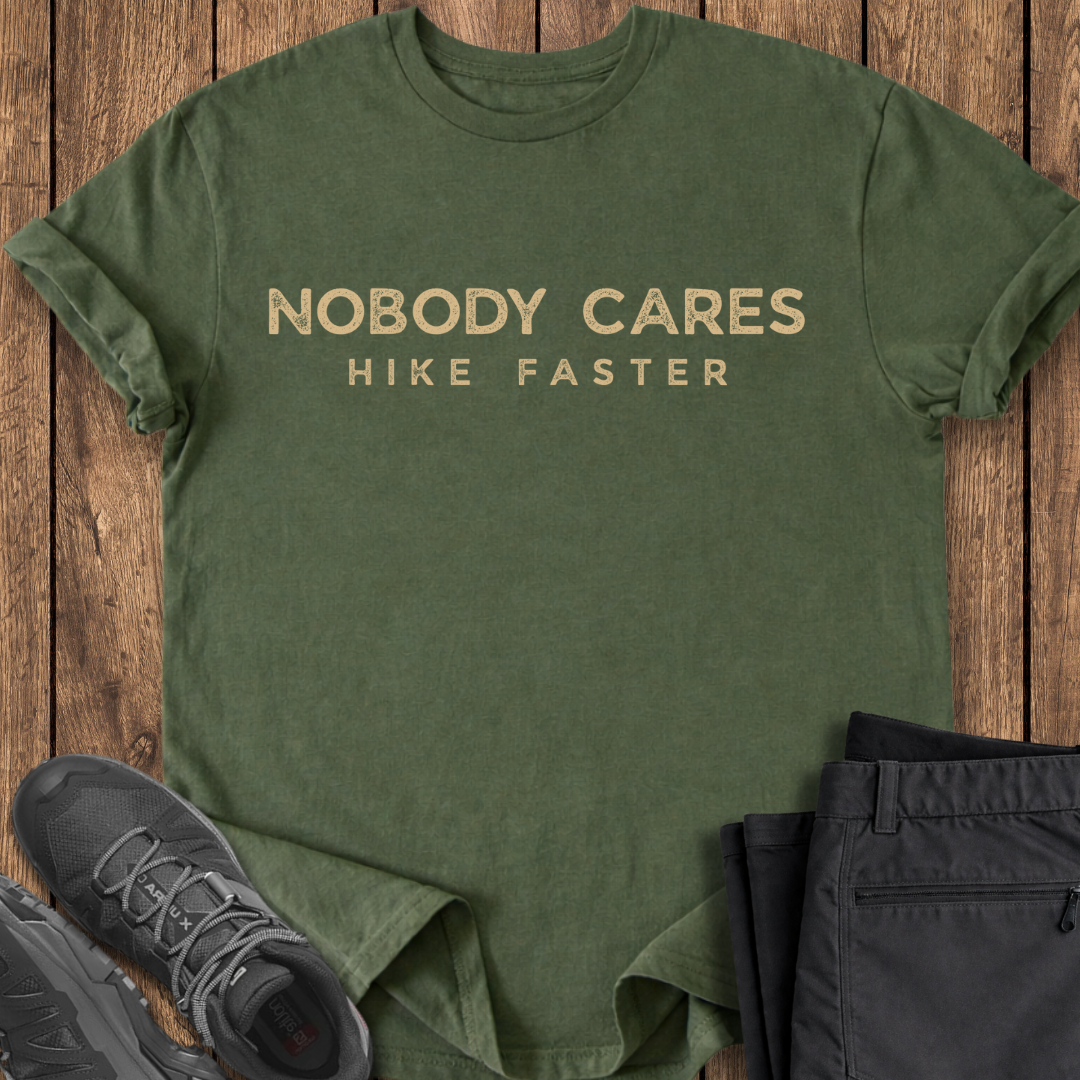 Green t-shirt with 'Nobody Cares Hike Faster' text on a wooden surface with hiking shoes and pants.