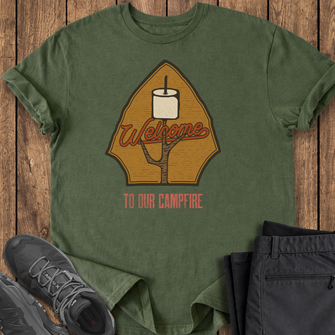 Green t-shirt with 'Welcome to our campfire' design on a wooden surface with shoes and pants. marshmellow marshmallow stick camping campfire