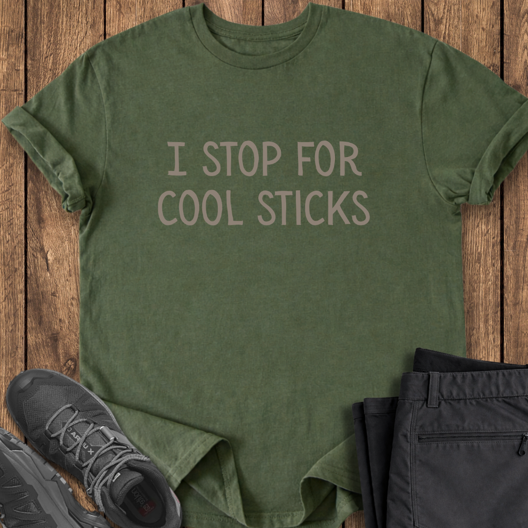 Green t-shirt with 'I Stop for Cool Sticks' text on a wooden surface with shoes and pants. funny hiking t shirt