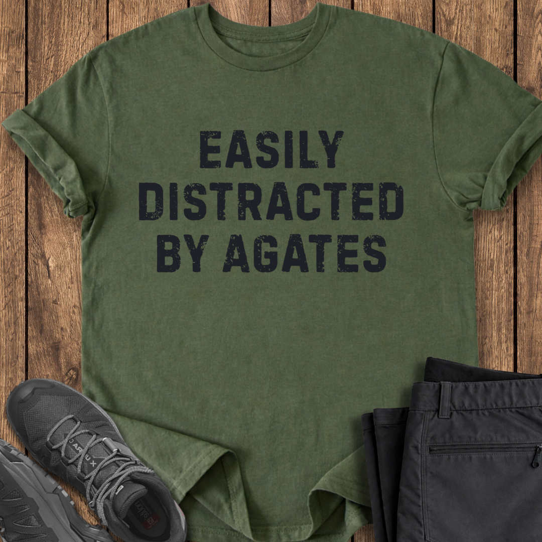 Green t-shirt with 'Easily Distracted by Agates' text on a wooden surface with shoes and pants.