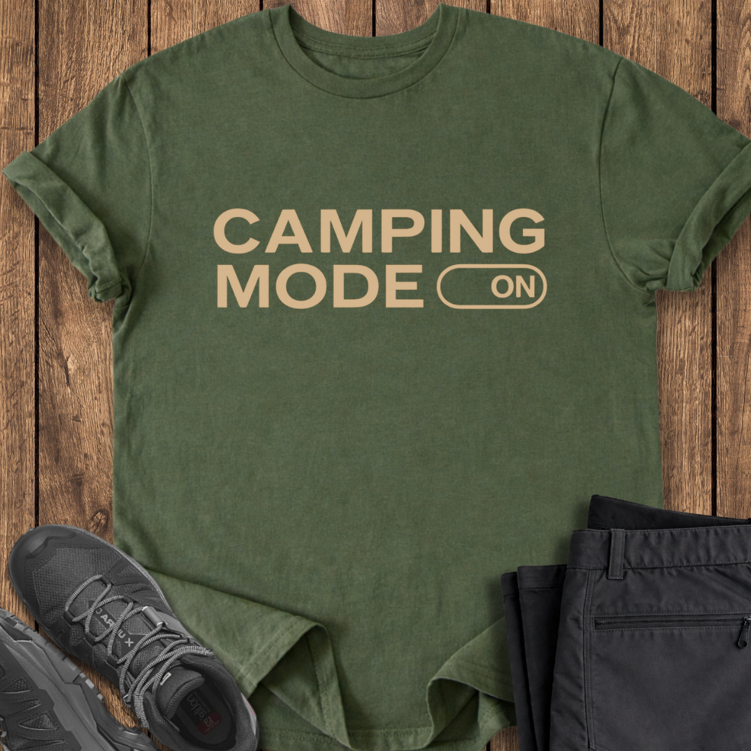 Green t-shirt with 'Camping Mode On' text on a wooden surface with shoes and pants.