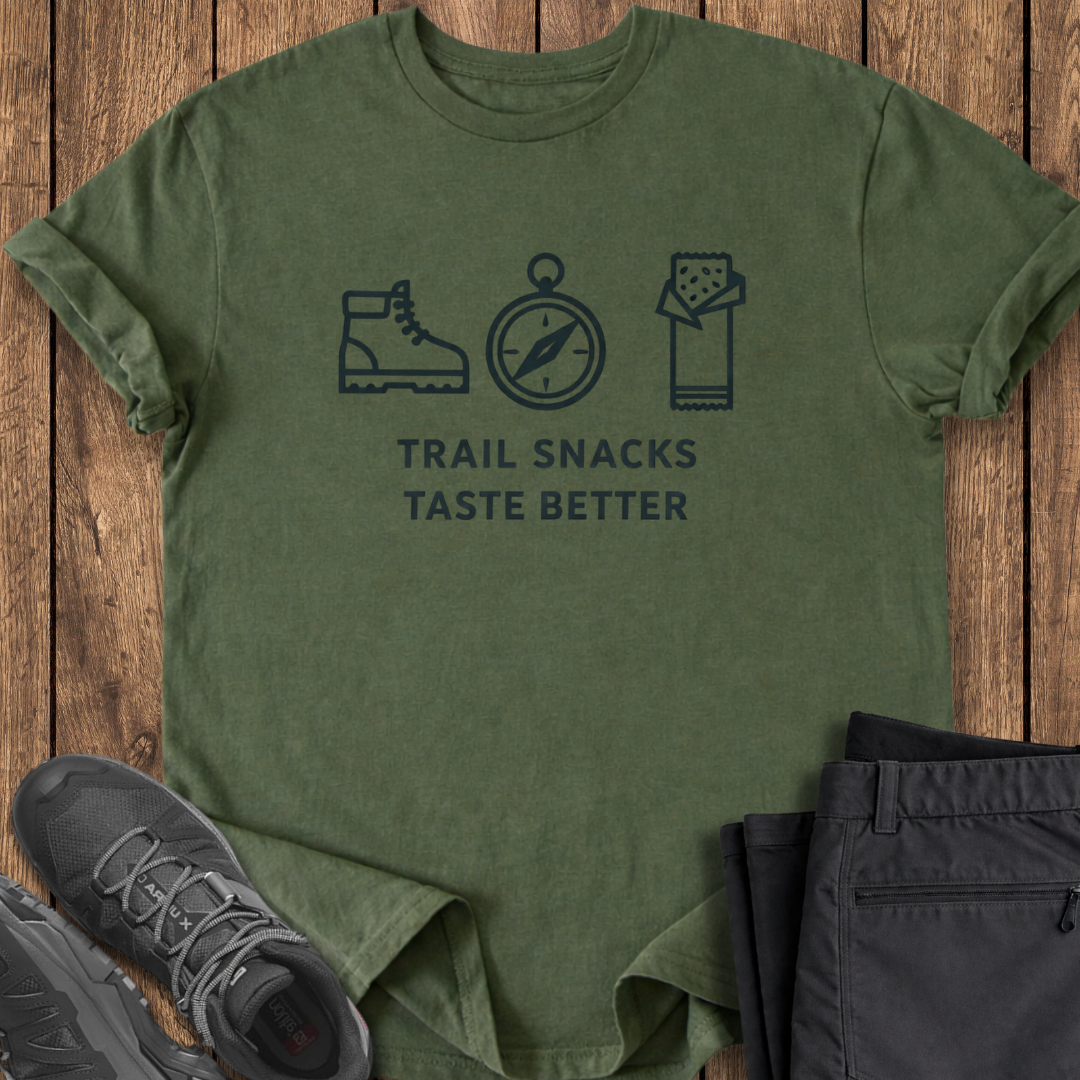 Green t-shirt with hiking icons and text on a wooden surface with shoes and pants. trail snacks taste better