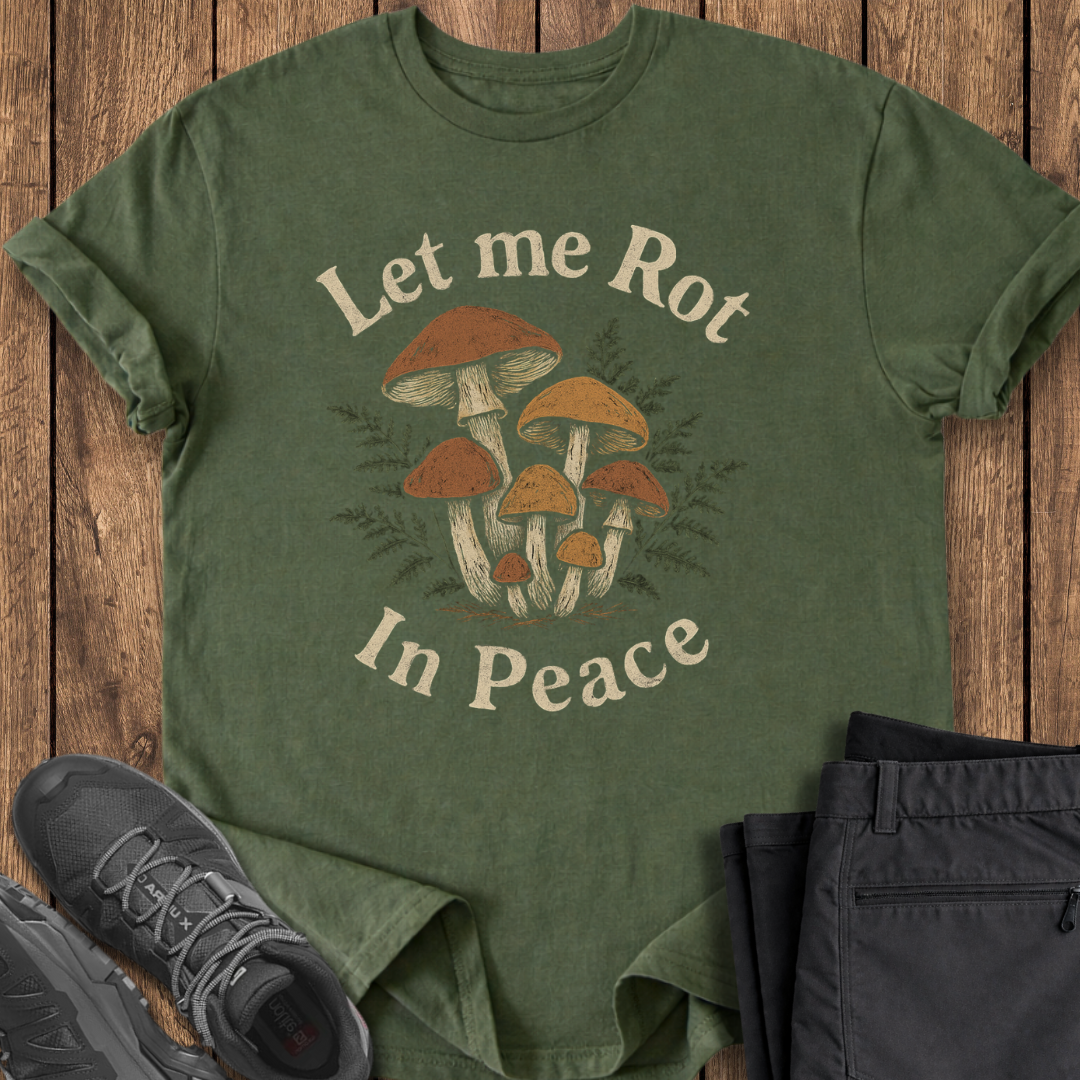 Green t-shirt with mushroom design and text 'Let me Rot In Peace' on a wooden surface with shoes and pants.