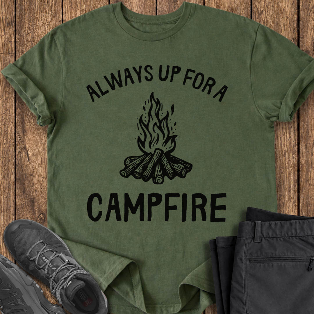 Green t-shirt with 'Always Up For a Campfire' design on a wooden surface with shoes and pants. camp camping t shirt