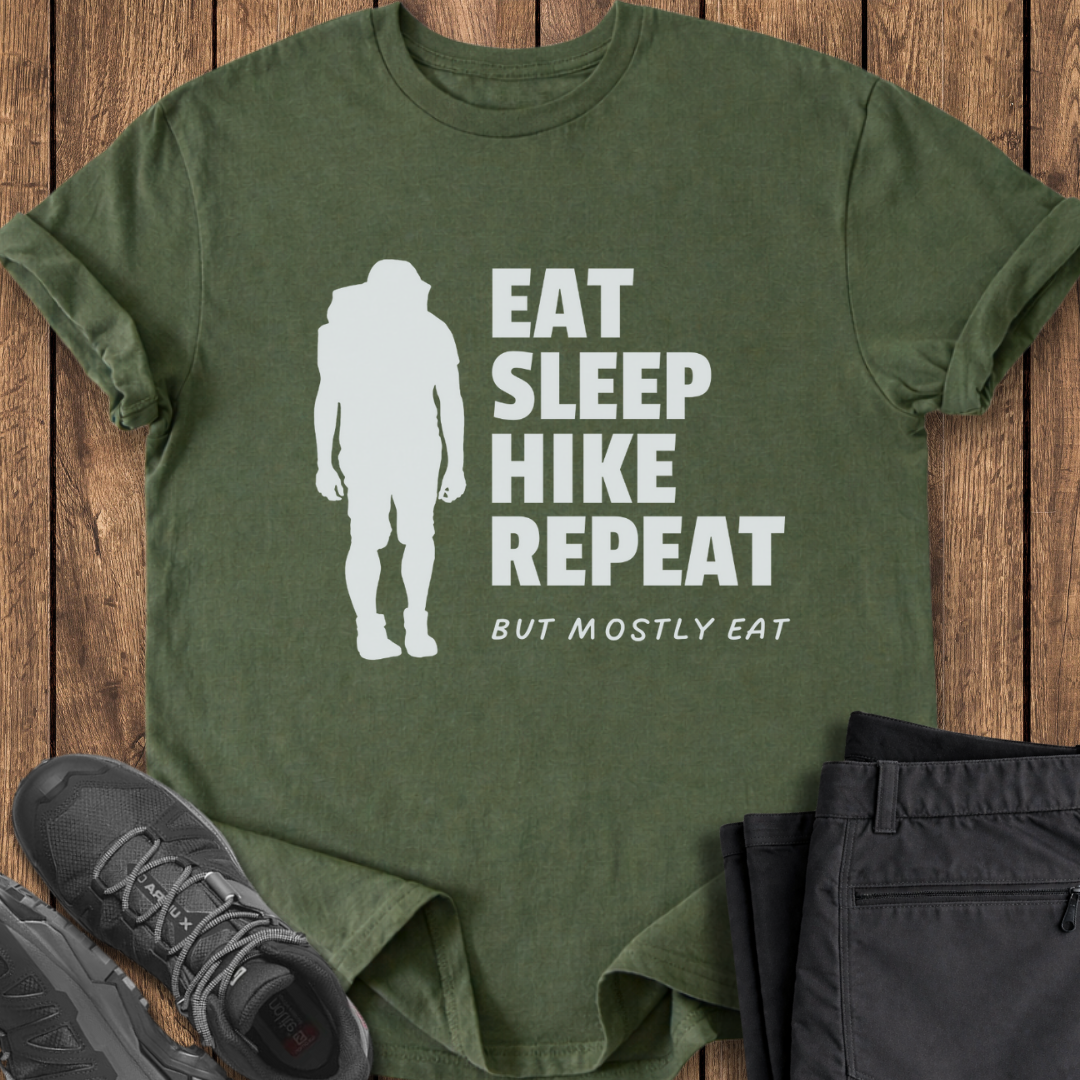 Green t-shirt with 'Eat Sleep Hike Repeat' text and silhouette on a wooden surface with hiking shoes and pants. funny hike t shirt