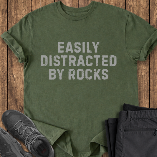 Green t-shirt with 'Easily Distracted By Rocks' text on a wooden surface with shoes and pants.