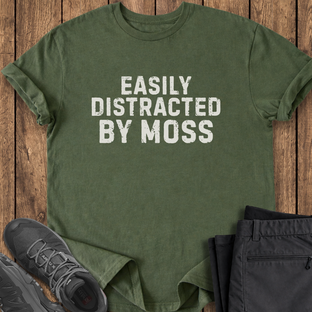 Green t-shirt with 'Easily Distracted by Moss' text on a wooden surface with shoes and pants.