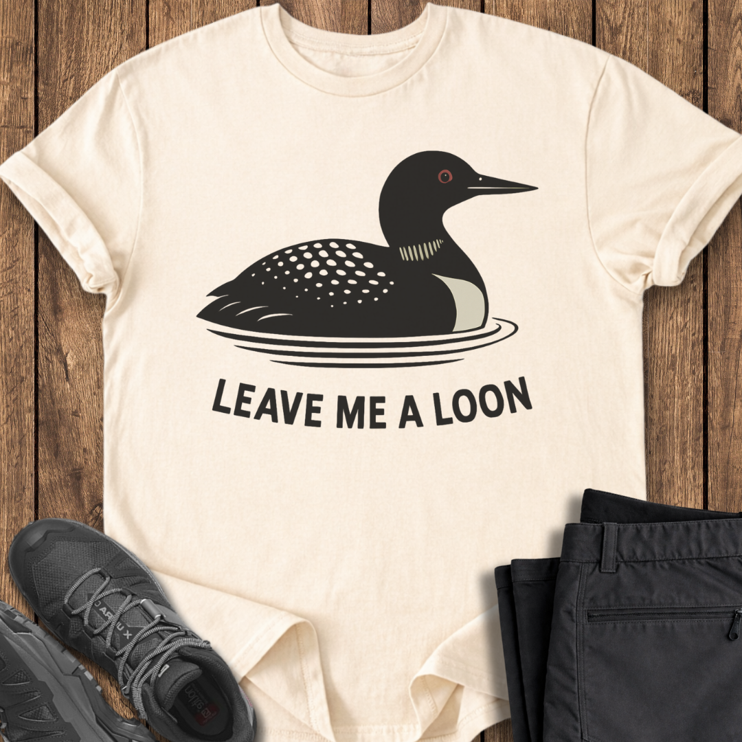 Beige t-shirt with a duck graphic and text 'Leave Me A Loon' on a wooden surface with shoes and pants.