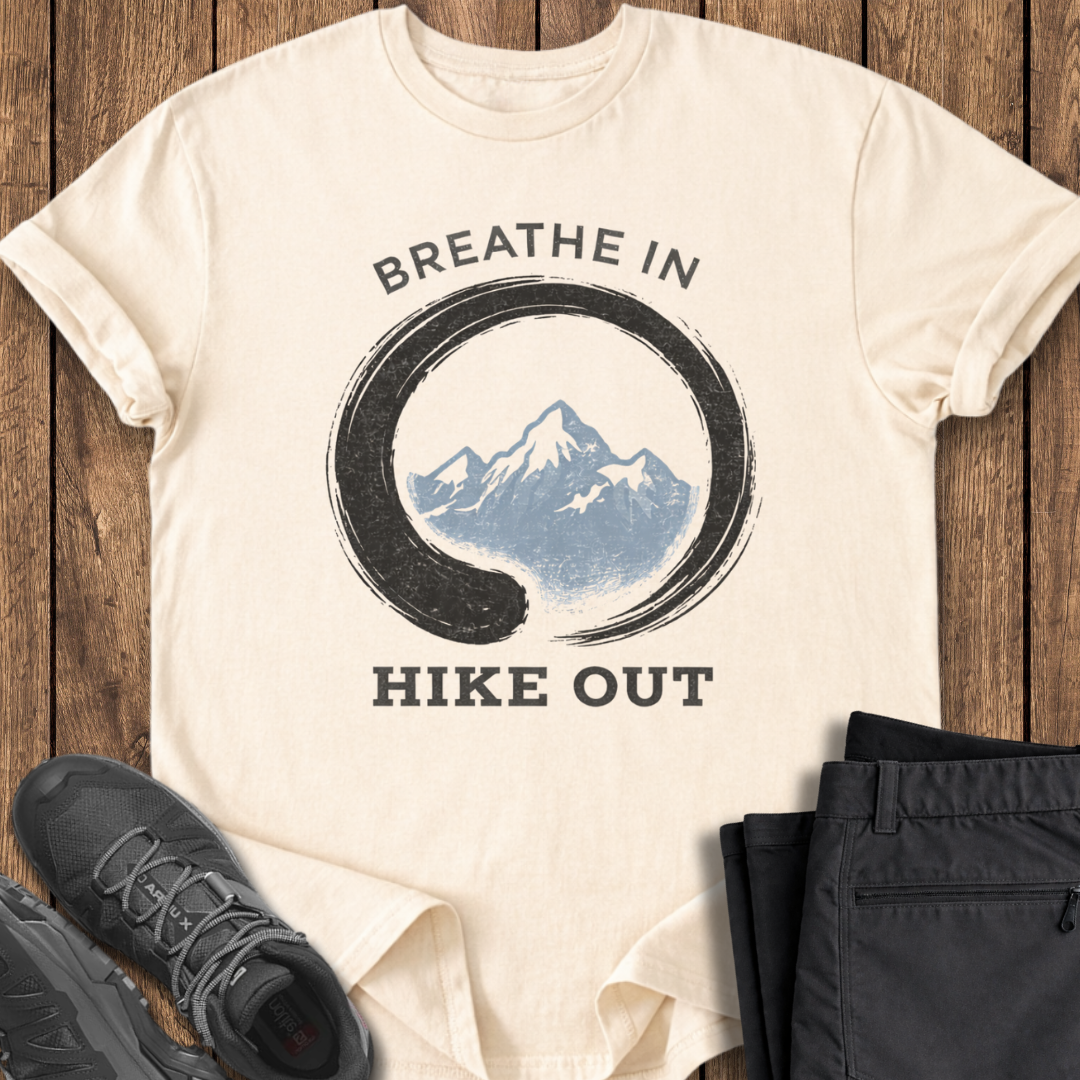 Beige t-shirt with mountain graphic and text 'Breathe In Hike Out' on a wooden surface with shoes and pants. mindful mountain t shirts