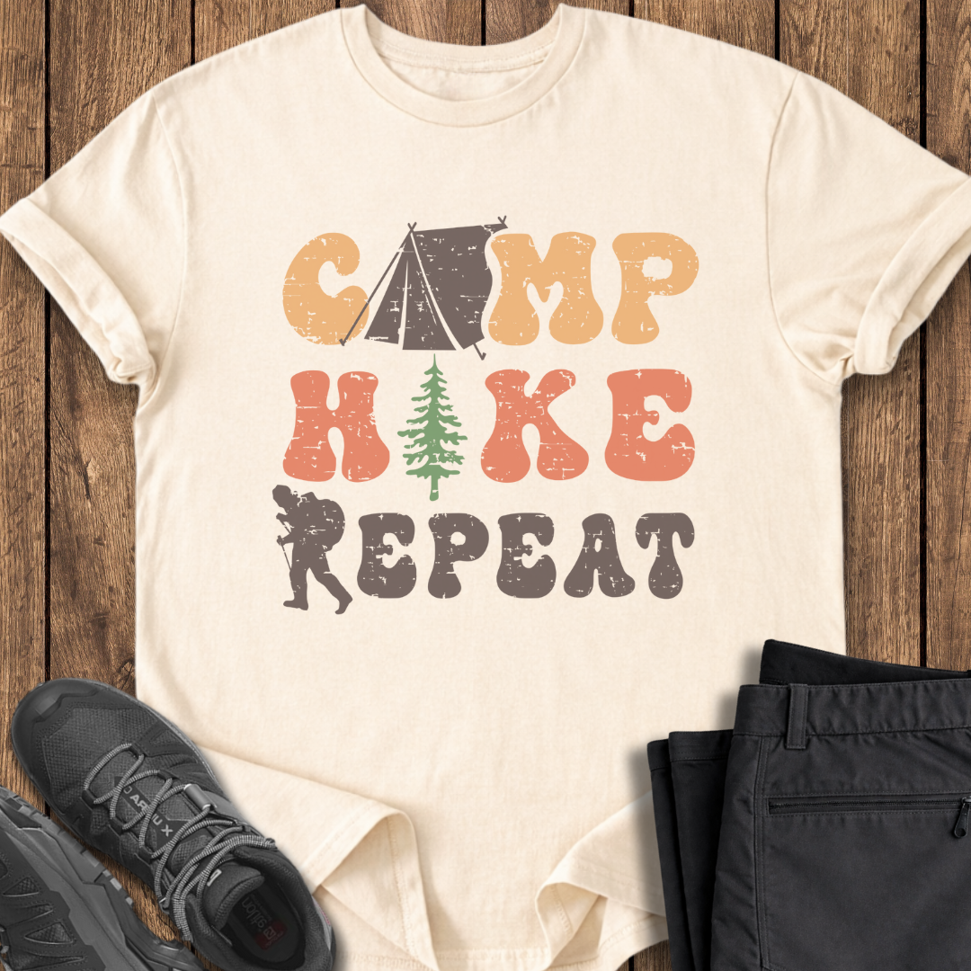 Beige t-shirt with 'Camp Hike Repeat' design on a wooden surface with shoes and pants.