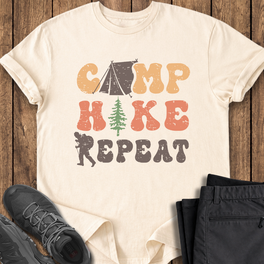 Beige t-shirt with 'Camp Hike Repeat' design on a wooden surface with shoes and pants.