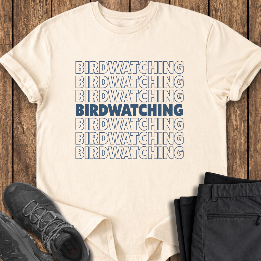 Beige t-shirt with 'BIRDWATCHING' text printed multiple times, paired with black pants and shoes on a wooden surface.