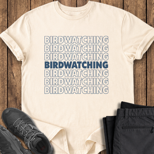 Beige t-shirt with 'BIRDWATCHING' text printed multiple times, paired with black pants and shoes on a wooden surface.