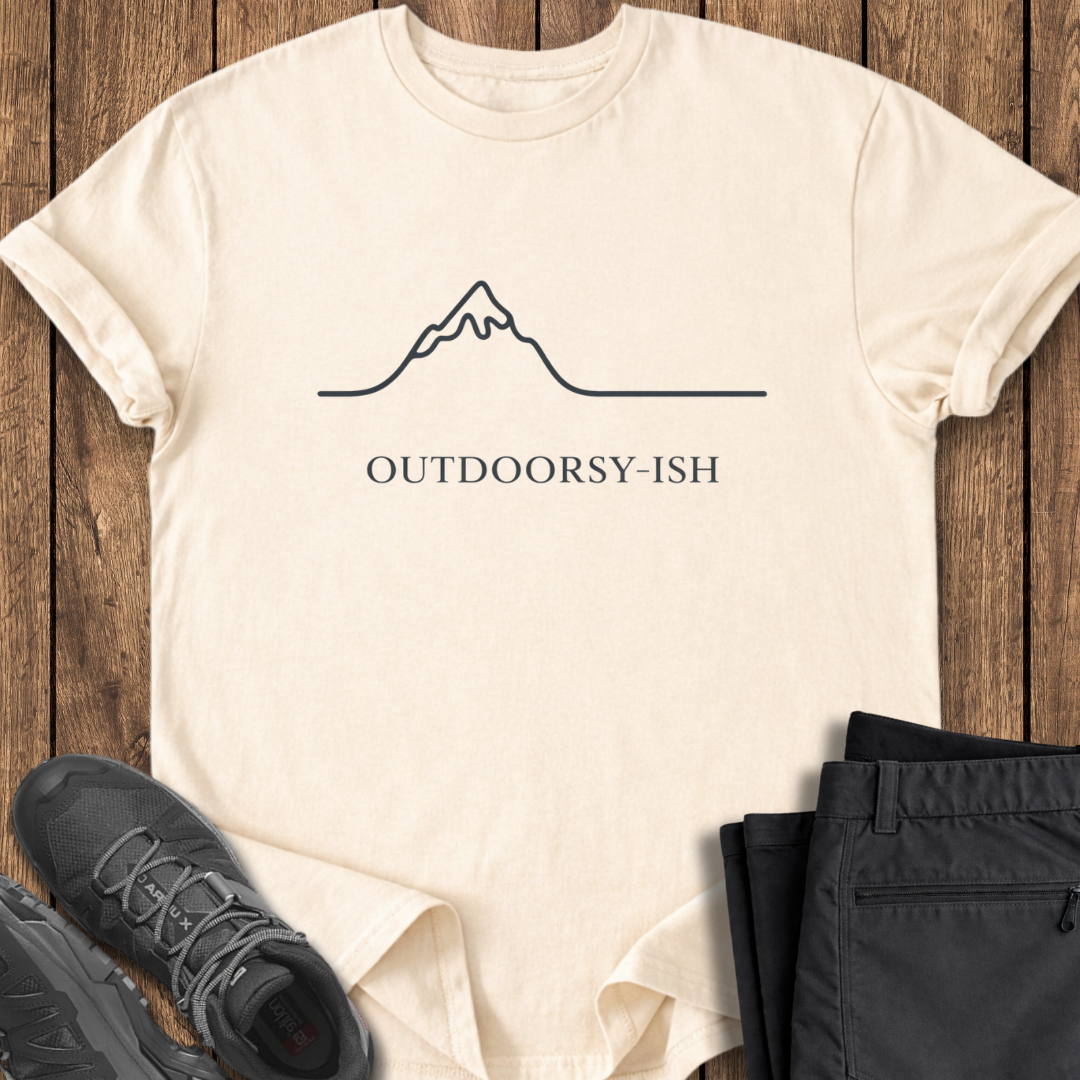 Beige t-shirt with 'OUTDOORSY-ISH' text and mountain graphic on a wooden surface, with black pants and shoes. funny mountain t shirt