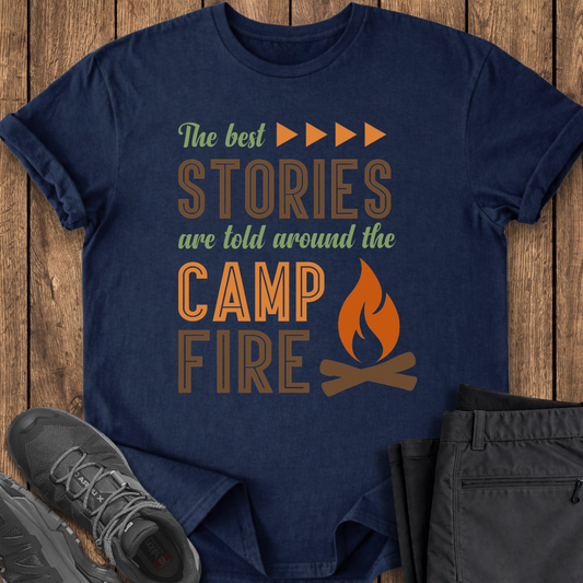 Navy blue t-shirt with 'The best stories are told around the campfire' text and graphic on a wooden surface with shoes and pants. camping outdoor shirt