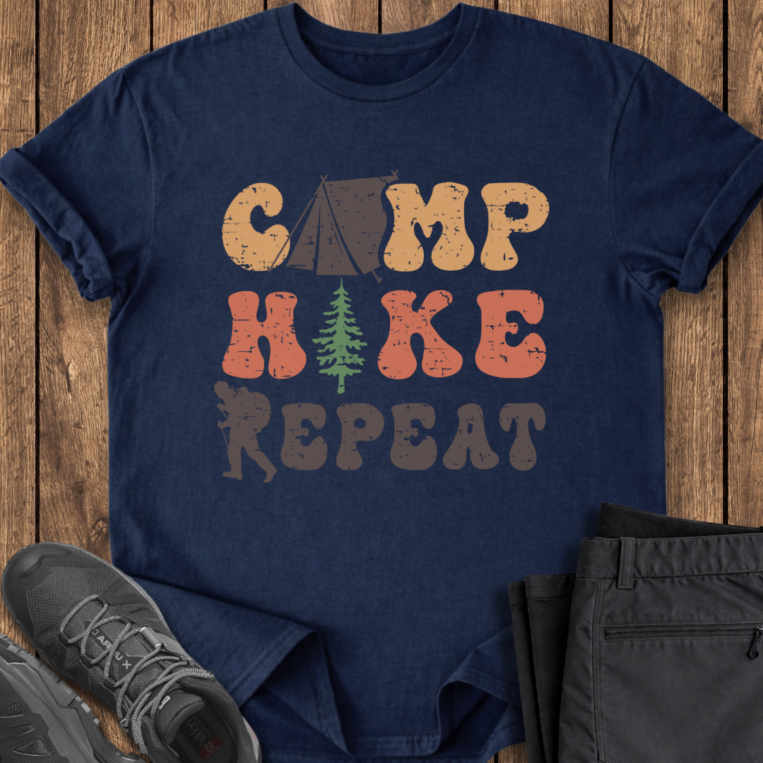 Navy blue t-shirt with 'Camp Hike Repeat' text and camping graphics on a wooden surface.