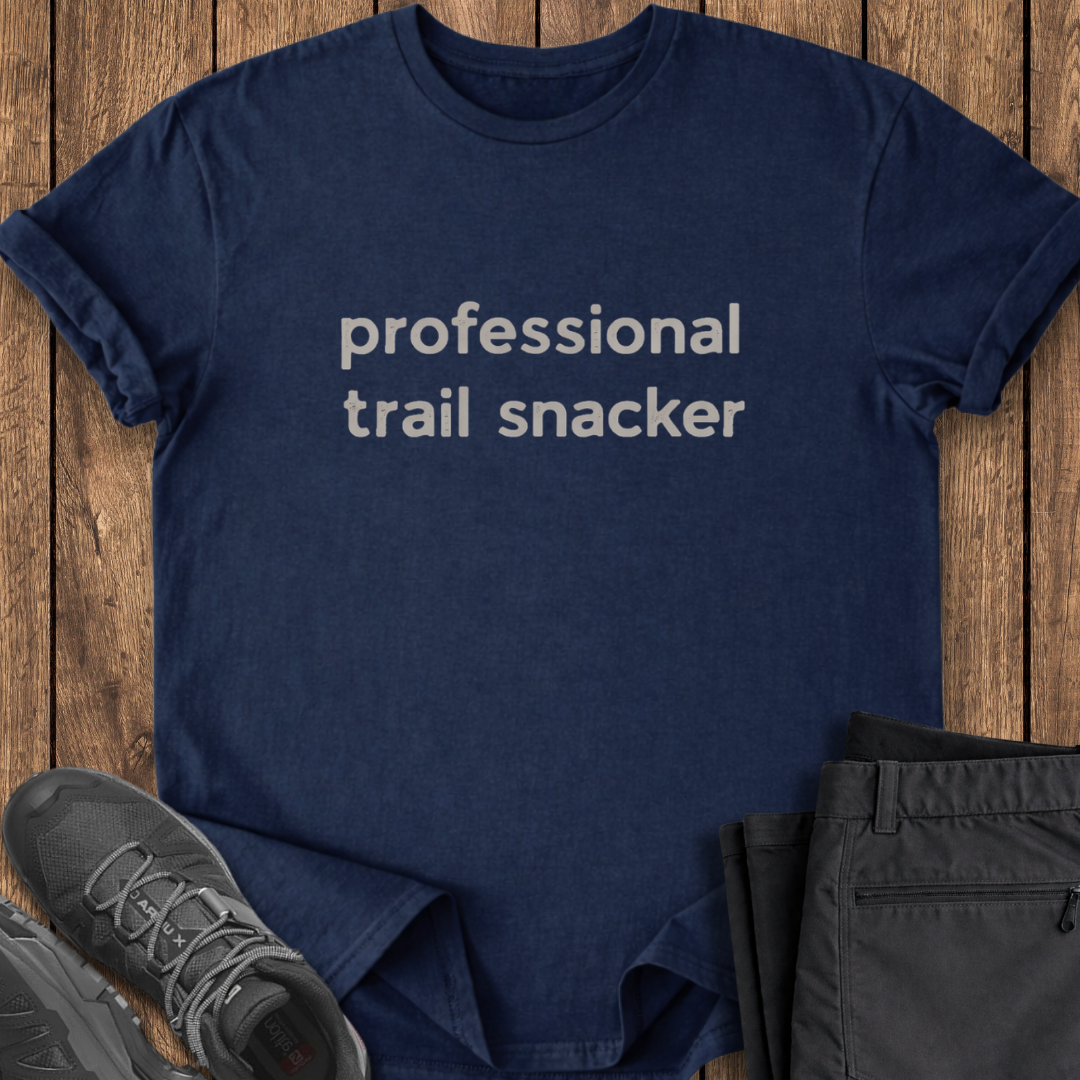 Navy blue t-shirt with 'professional trail snacker' text on a wooden surface with shoes and shorts. funny hike t shirt
