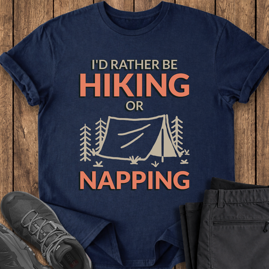 Navy blue t-shirt with 'I'd Rather Be Hiking or Napping' text and tent graphic on a wooden surface with shoes and pants. funny hiking camping t shirt