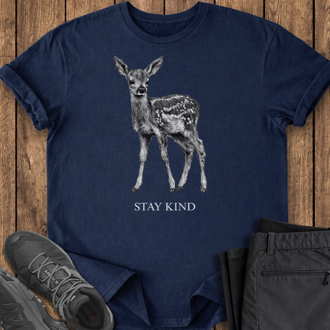 Navy blue t-shirt with a deer graphic and 'Stay Kind' text on a wooden surface, with shoes and pants partially visible.