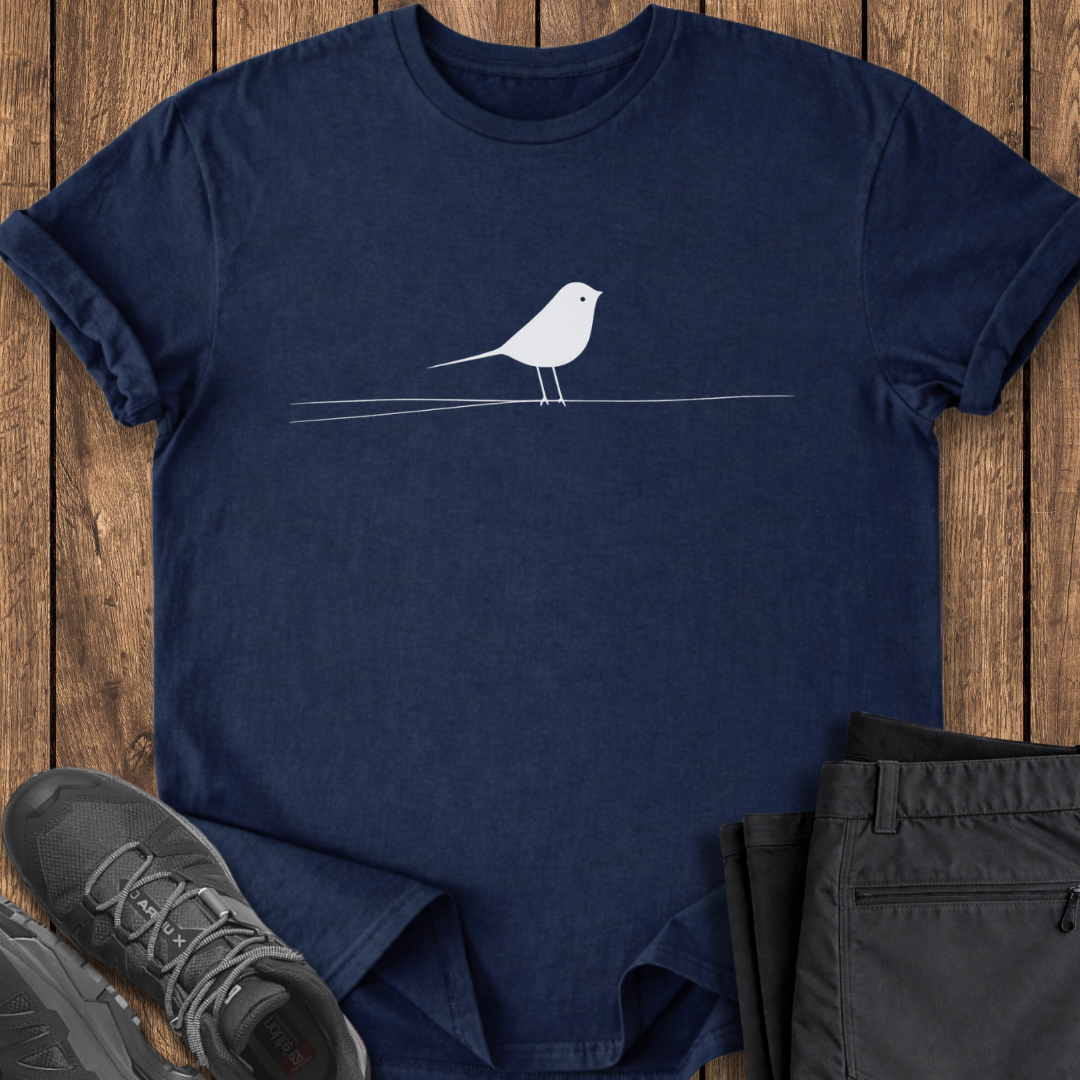 Navy blue t-shirt with a white bird design on a wooden surface, surrounded by gray shoes and black pants.