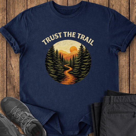 Navy blue t-shirt with 'Trust the Trail' graphic on a wooden surface with shoes and pants. hiking trail through the pine forest sunset hiking t shirt
