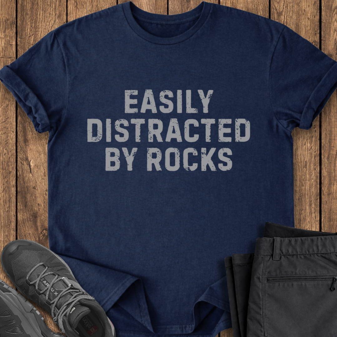 Navy blue t-shirt with 'Easily Distracted By Rocks' text on a wooden surface with shoes and pants.