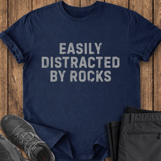 Navy blue t-shirt with 'Easily Distracted By Rocks' text on a wooden surface with shoes and pants.
