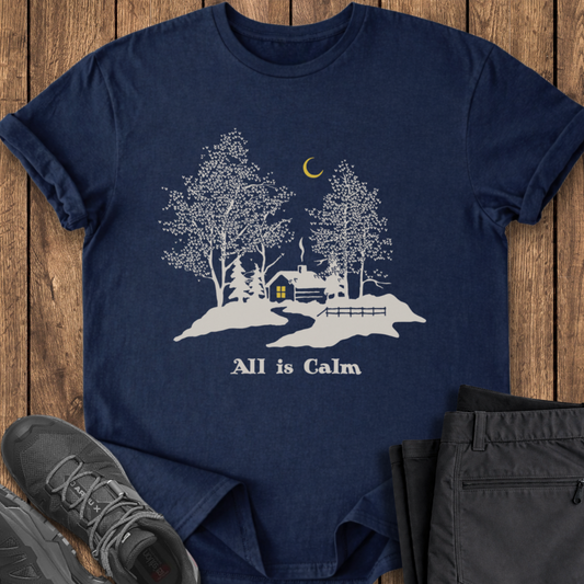 Navy blue t-shirt with a white graphic and 'All is Calm' text on a wooden surface, with shoes and pants for scale. winter snow cabin 