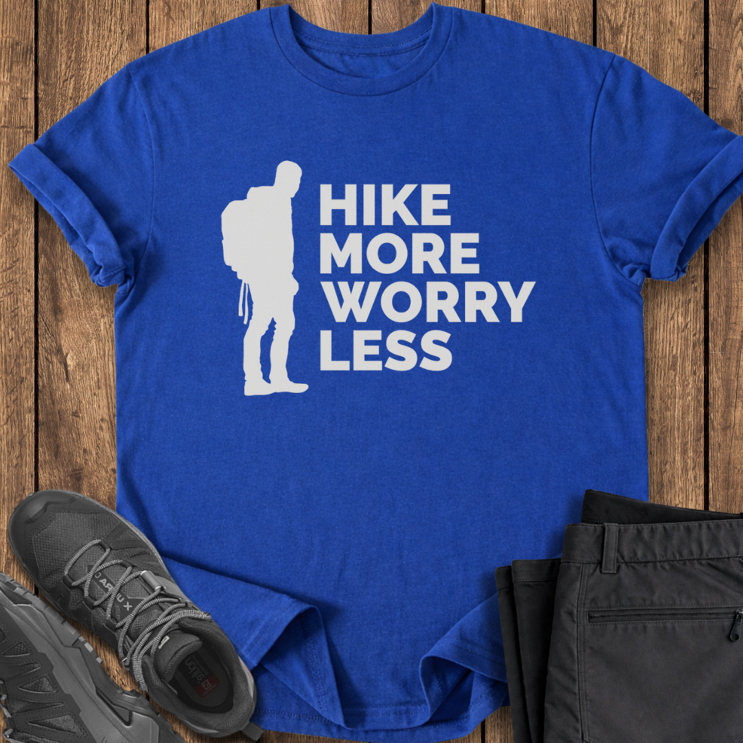 Blue t-shirt with 'Hike More Worry Less' design on a wooden surface with hiking shoes and pants.