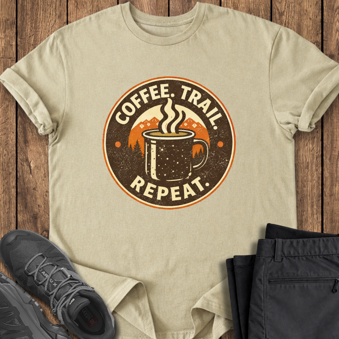 Beige t-shirt with 'Coffee. Trail. Repeat.' graphic on a wooden surface with shoes and pants. coffee hiking t shirts