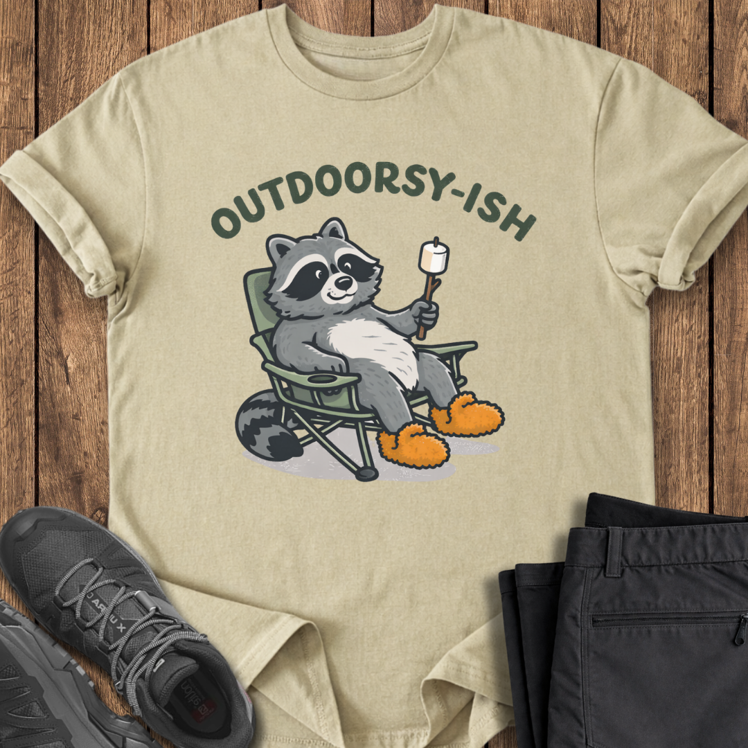 Beige t-shirt with raccoon graphic and 'Outdoorsy-ish' text on a wooden surface with shoes and pants. smore outdoor hiking t shirt