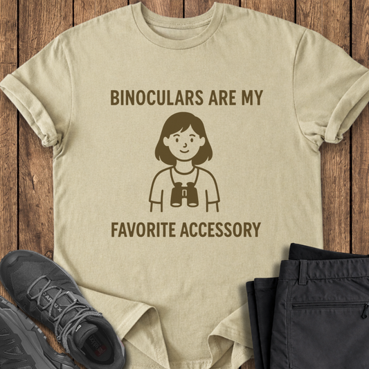 Beige t-shirt with a graphic and text 'Binoculars are my favorite accessory' on a wooden surface with shoes and pants.