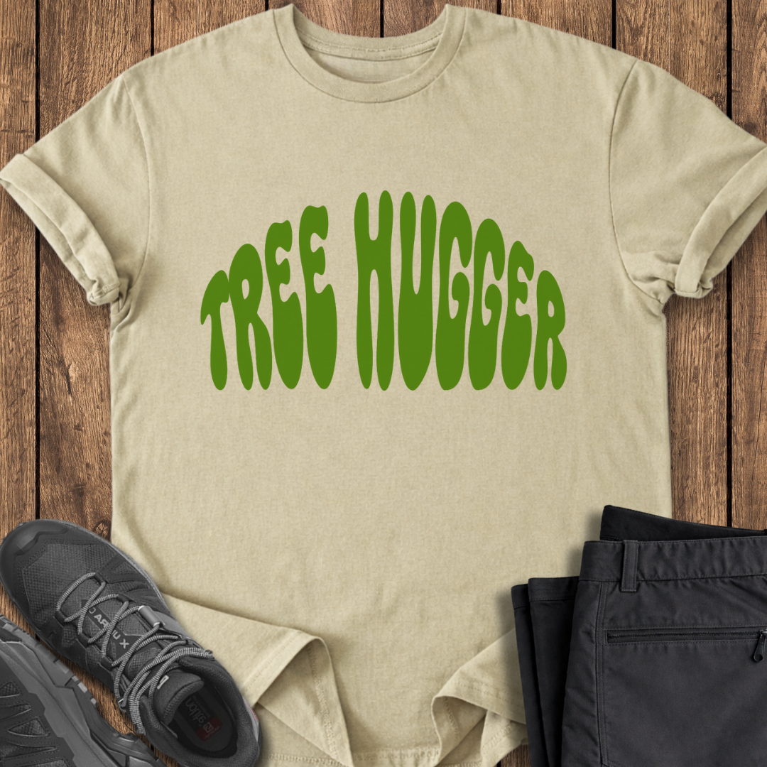 Beige t-shirt with 'TREE HUGGER' text in green on a wooden surface with black sneakers and pants.