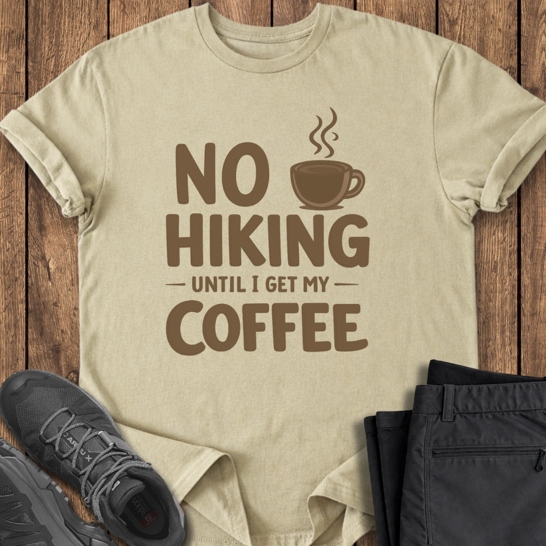 Beige t-shirt with 'No Hiking Until I Get My Coffee' text and graphic on a wooden surface with shoes and pants. funny hiking t shirt