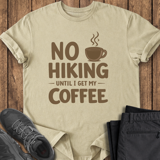 Beige t-shirt with 'No Hiking Until I Get My Coffee' text and graphic on a wooden surface with shoes and pants. funny hiking t shirt