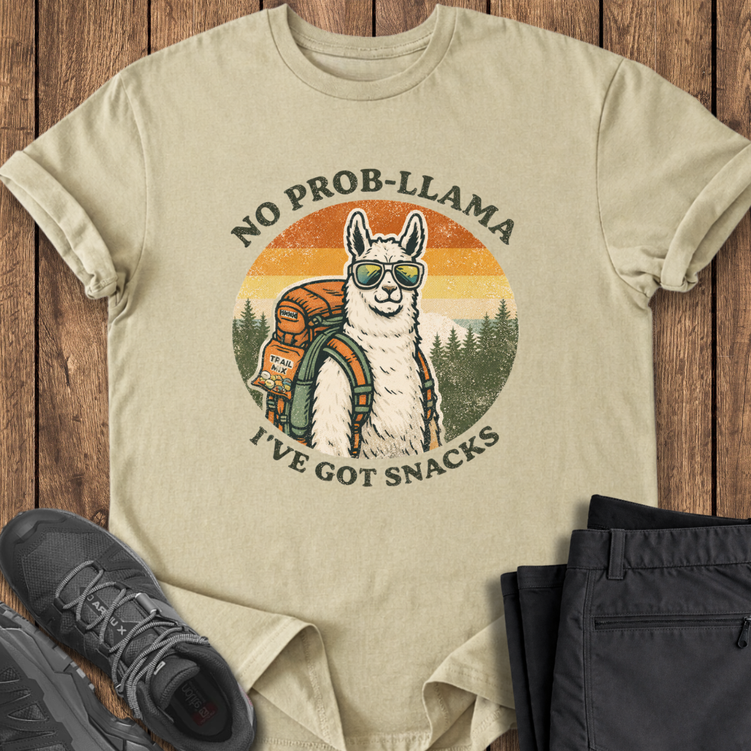Beige t-shirt with a llama graphic and text 'No Prob-Llama, I've Got Snacks' on a wooden surface with shoes and pants. funny hiking t shirt